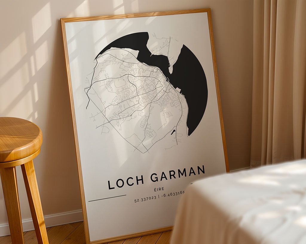 The FN Prints Wexford City Map, a minimalist black-on-white framed poster featuring coordinates "52.337023" and "-6.433166," rests against a wall with sunlight casting shadows beside a nearby wooden stool and curtain, highlighting its high-quality design.