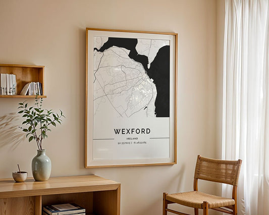 FN Prints' framed Wexford City Map, in high-quality print, adorns a beige wall with its GPS coordinates below. A wooden shelf with books and a vase of green branches complements the minimalist design to the left, while a wooden chair rests under a sheer white curtain on the right.