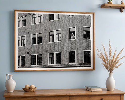 Window Watcher" by FN Prints, a framed black and white urban photo of a brick building, hangs on a light blue wall above a wooden sideboard decorated with vases, a bowl, and a shelf holding books and a small plant.
