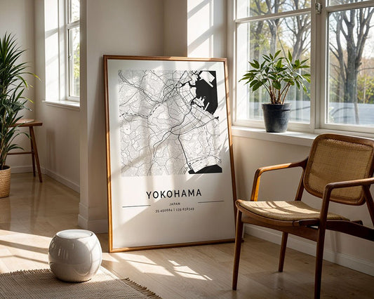 A framed FN Prints Yokohama City Map leans against a window in a sunlit room, where a wicker chair, potted plant, and small white stool adorn the wooden floor, crafting an inviting atmosphere.