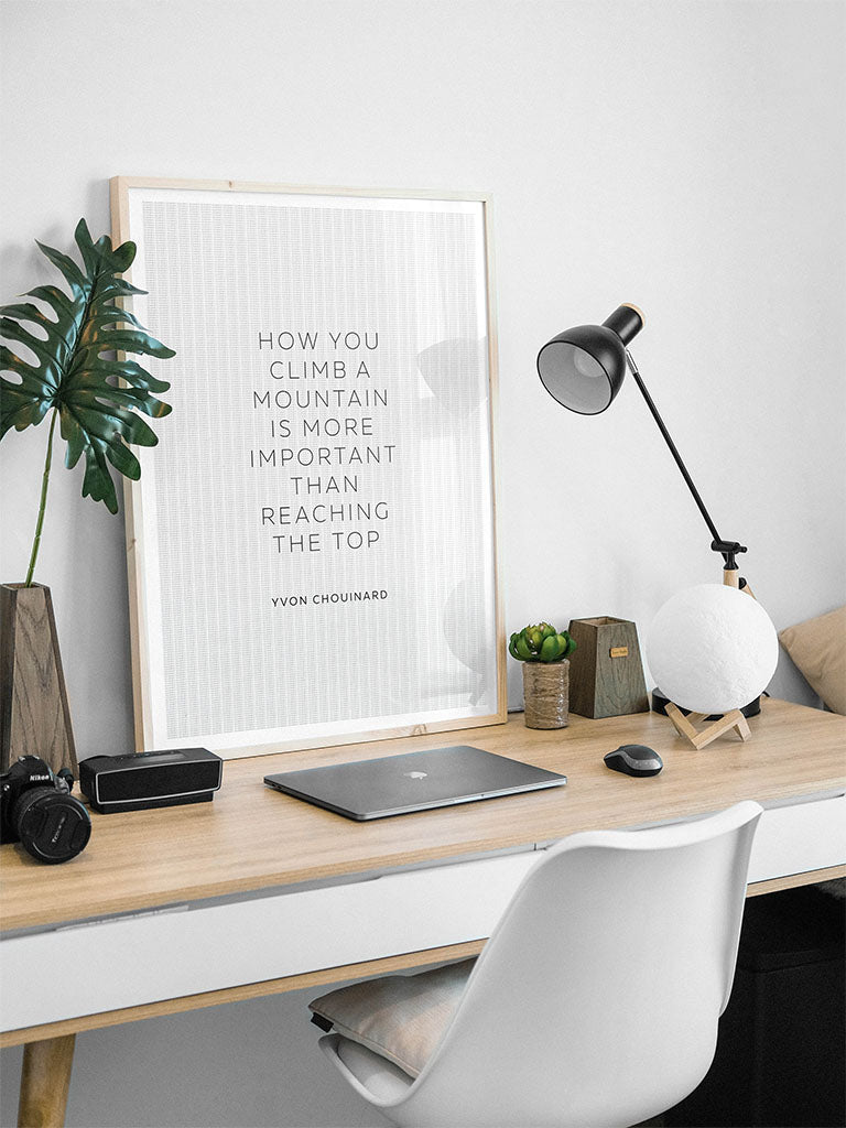 A minimalist desk setup features a white chair, laptop, camera, lamp, small plants, and the FN Prints "Yvon Chouinard - How You Climb a Mountain" art print as motivational decor.