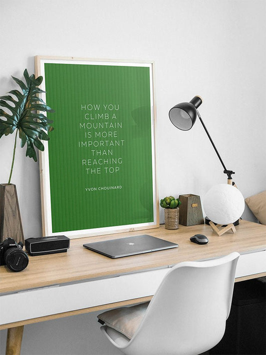 A minimal desk setup features a white chair, wooden desk, laptop, lamp, small plants, camera, and a framed FN Prints "Yvon Chouinard - How You Climb a Mountain" art print for motivation.