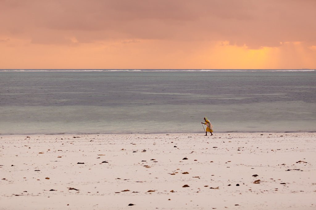 A solitary figure with a staff strolls along an expansive, deserted Zanzibar beach dotted with rocks. The sky above is a blend of orange and pink tones, indicating either an early morning or late evening sunset by the tranquil sea—a genuine display of cultural authenticity in this "Zanzibari Woman" sunset photograph by FN Prints.