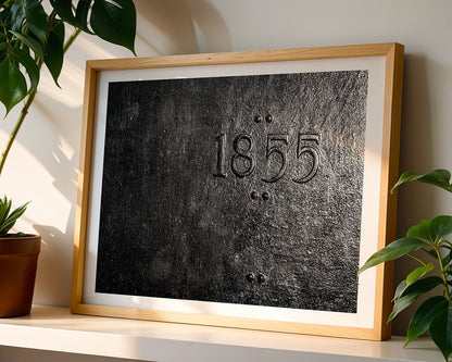 The "1855" by FN Prints, a framed artwork featuring a textured black surface with the year embossed at its center, showcases industrial elegance. When placed on a white shelf, it elevates any art collection, beautifully complemented by surrounding green plants and sunlight creating gentle shadows across the scene.