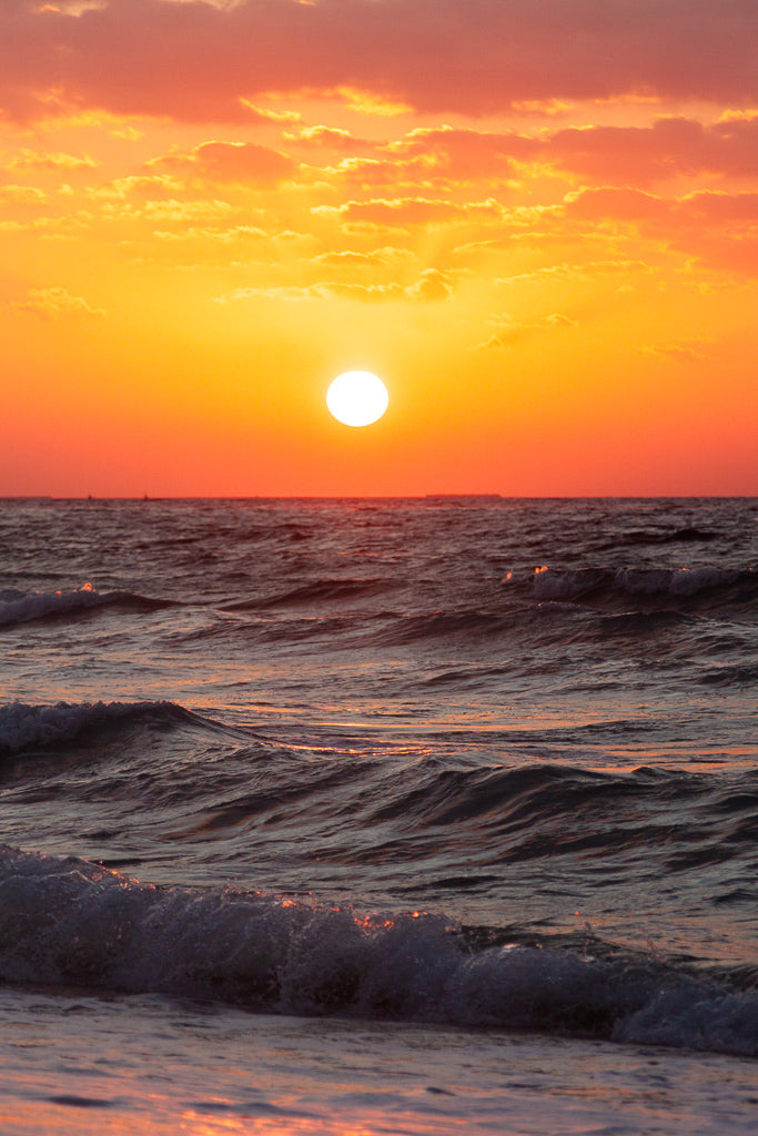 Experience the breathtaking beauty of a Seaside Sunset with this exquisite photographic print from FN Prints. The sun, appearing as a bright glowing orb, is partially veiled by orange and pink-tinged clouds, casting a warm golden light over the gently rippling waves below. This stunning scene captures the serene ocean horizon in perfect detail.