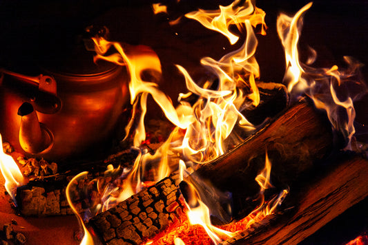 A close-up of *The Fiery Teapot* by FN Prints, near a campfire with bright, dancing flames and burning wooden logs. To the left, the vintage teapot is placed near the edge of the fire, partially engulfed by the warm glow of the flames. The scene radiates rustic charm and a sense of warmth and coziness.