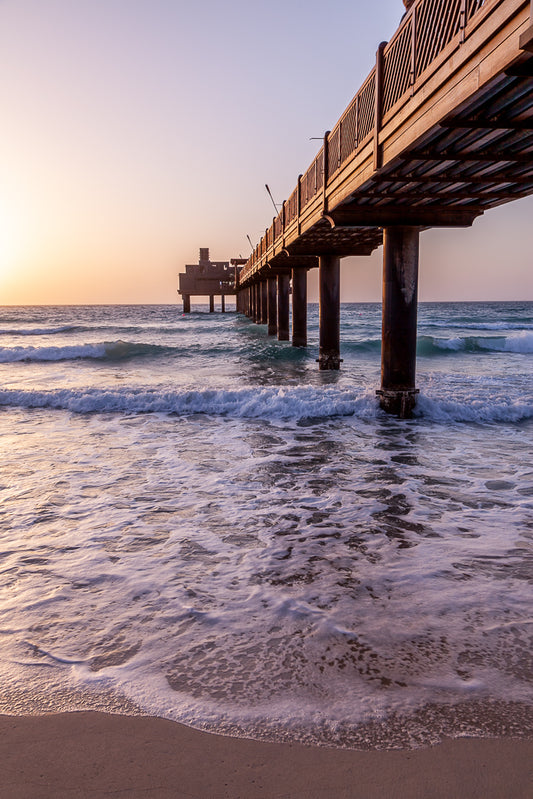 The Pier" by FN Prints captures a wooden structure extending over waves gently crashing on the beach at sunset—ideal for coastal wall art. The beams of the pier cast shadows over the water, and the sky transforms from orange near the horizon to a light purple higher up, encapsulating a beautiful golden hour sunset.