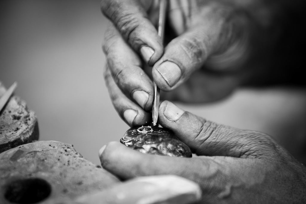 A close-up, black-and-white image captures a pair of hands meticulously working on what appears to be handmade jewelry. One hand holds the object while the other uses a small tool to make precise adjustments, showcasing the intricate hand-crafting skills involved. This scene is beautifully illustrated in FN Prints' "Kandy Jeweller's Hands.