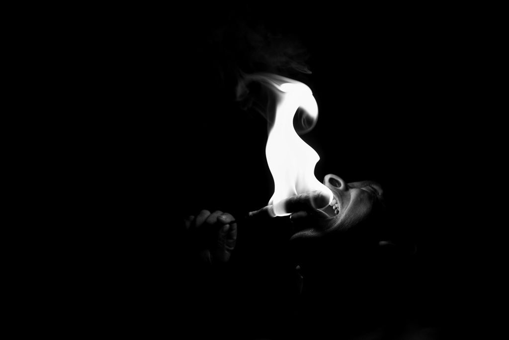 In FN Prints' black and white photograph "Fire Eater," the performer showcases their fiery skill in a dimly lit environment. The brilliant flame stands out sharply against the dark backdrop, casting light on parts of their face and the tip of the torch held near their mouth.