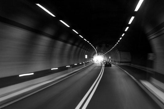 Introducing "Tunnel Vision" by FN Prints: This black-and-white photograph vividly captures a car driving through a well-lit, two-lane tunnel with solid double lines. The slight blur of the scene conveys dynamic motion and encapsulates the essence of urban life, with the light at the tunnel's end visible in the distance.