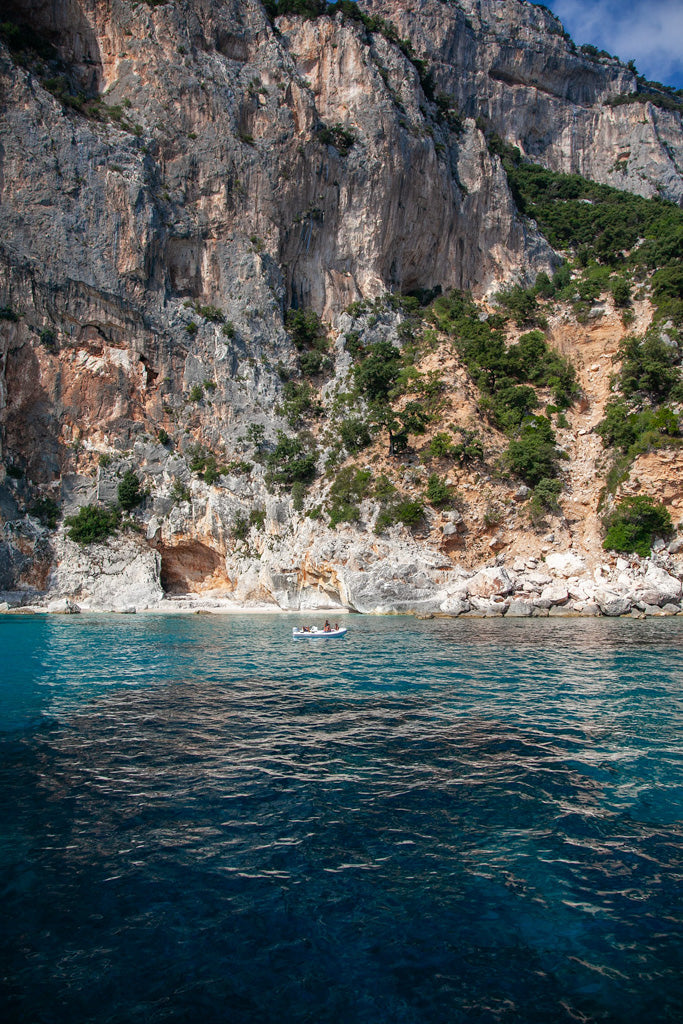 A small boat floats on clear turquoise waters near the rugged cliffs of the Sardinian Coast, which are covered with lush green foliage. The cliff rises steeply, showcasing textured rock faces. The calm waters reflect the blue sky, creating a striking contrast with the rugged terrain captured in this beautiful coastal photograph by FN Prints.