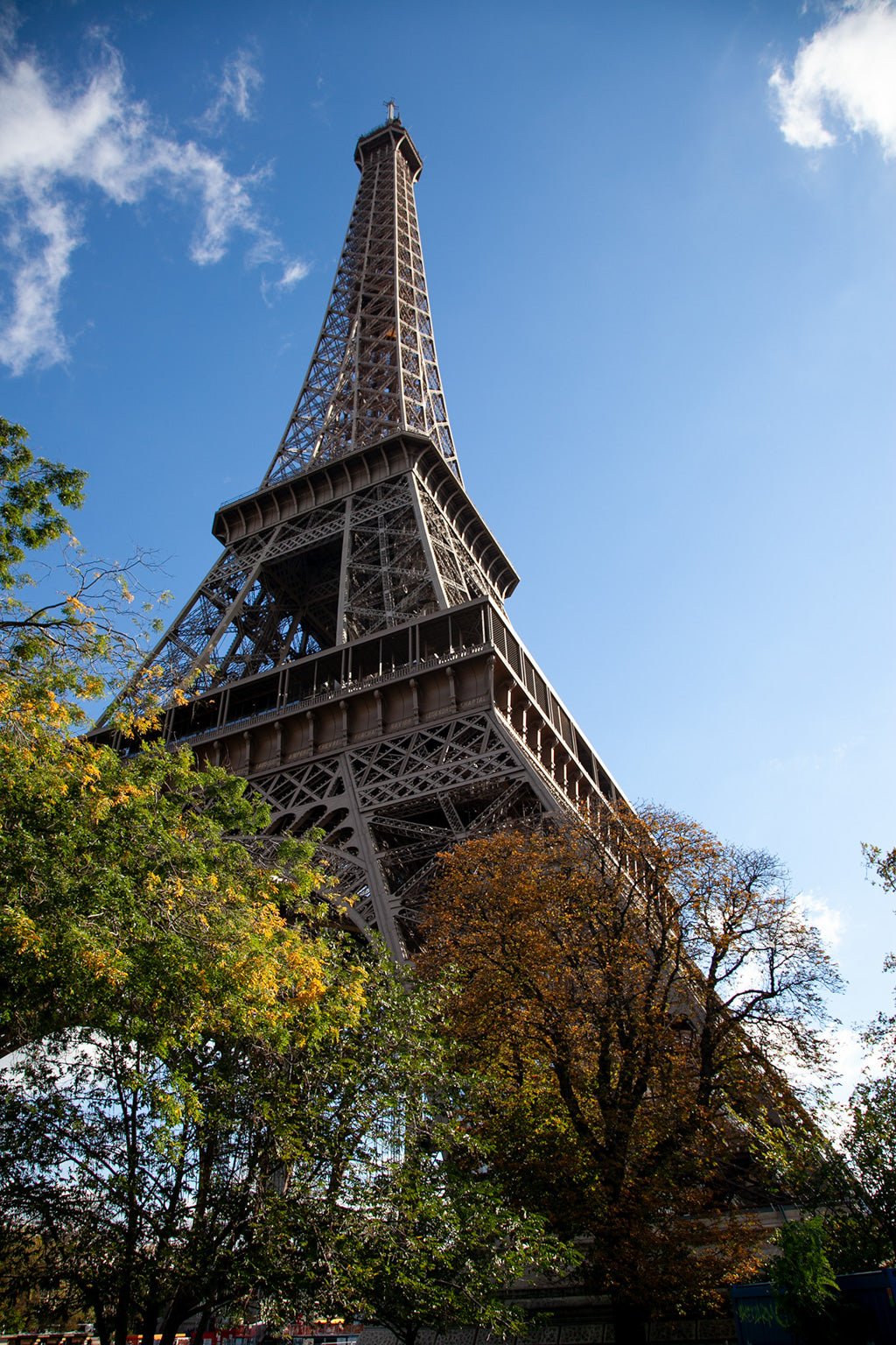 The Eiffel Tower: Part II Colour by FN Prints showcases the iconic Eiffel Tower in Paris, France, viewed from below against a clear blue sky. Trees adorned with autumn leaves surround the base of the tower, infusing vibrant colors into the scene. This picturesque view is transformed into an exquisite art print that perfectly captures the essence of Parisian charm.