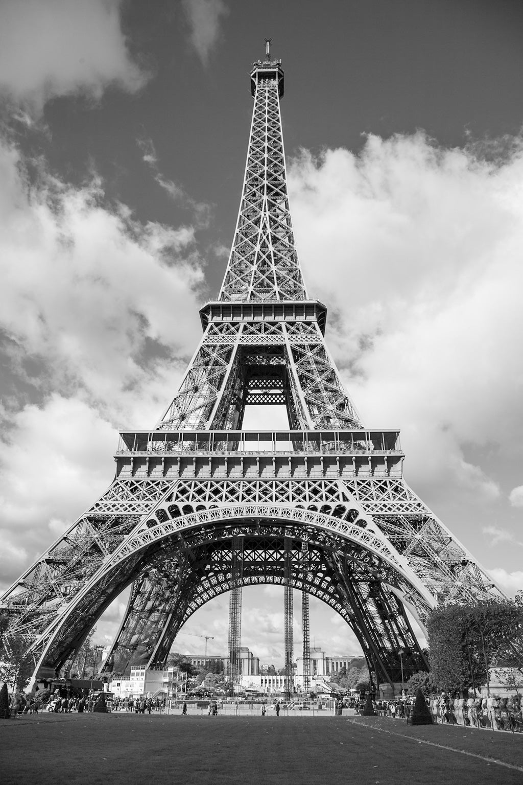 The Eiffel Tower: Part III Black & White by FN Prints is a striking black-and-white photograph that captures the iconic steel lattice structure of the Eiffel Tower from a low angle against a partly cloudy sky. The image includes trees and park areas at the base, where people are gathered, adding to the overall magnificence of this stunning piece of architectural artwork.