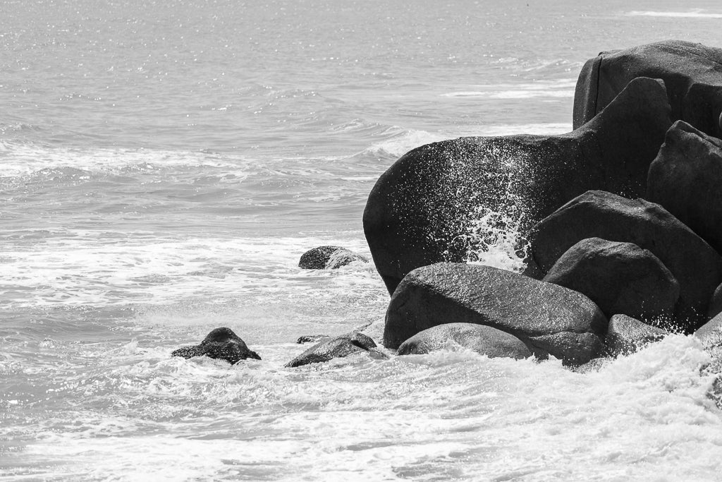 The Black Rocks print by FN Prints captures the intricate beauty of ocean waves crashing against large, dark rocks on the shore. Water splashes upon impact, creating a misty spray. This striking coastal scene showcases nature's raw power as the moderately rough sea meets the horizon in the background.