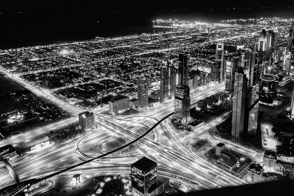 An aerial view of a sprawling cityscape at night, featuring the illuminated network of highways and roads, including the iconic Sheikh Zayed Road, is beautifully captured in "Urban Echoes" by FN Prints. Tall skyscrapers and buildings are scattered throughout, with bright lights outlining the urban grid. This stunning piece showcases modern infrastructure and dense urban development in remarkable detail.