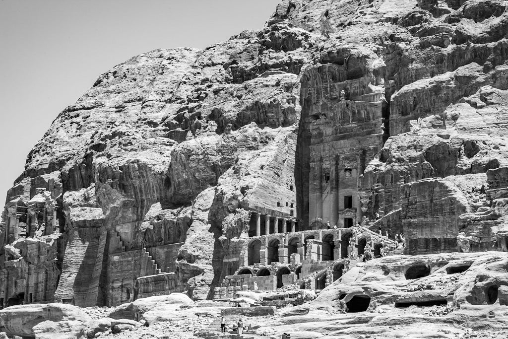 Black and white photo of ancient rock-cut architecture in Petra, Jordan. The structures, including the "Urn Tomb Petra, Jordan" from FN Prints, are carved into a rugged cliff, featuring columns, facades, and cave-like openings. The detailed stonework contrasts with the natural ruggedness of the surrounding rock face.