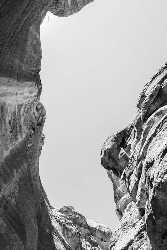 Through The Ravine by FN Prints: A black and white photo showcasing the towering, rugged rock formations of Wadi Rum canyon from below. The intricate textures and layers of the rocks create dramatic contrasts, emphasizing the minimalist aesthetics and raw natural beauty against a clear sky.