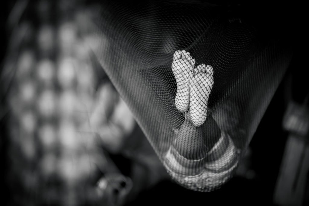 Suspended Serenity by FN Prints captures a black and white photograph of a small child's feet through netting, creating a striking textured pattern. The background is softly blurred and abstract, highlighting the delicate and tender quality of the child's feet, reminiscent of relaxation in a hammock.