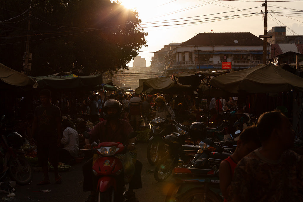 The scene depicted in "Sunset Market" by FN Prints captures the vibrancy of an outdoor market at sunset in Phnom Penh. People walk along the street lined with stalls beneath canopies, where motorbikes are parked nearby. The warm glow of the Cambodian sunset creates a play of shadows and highlights, beautifully accentuating the cultural richness showcased by this print.