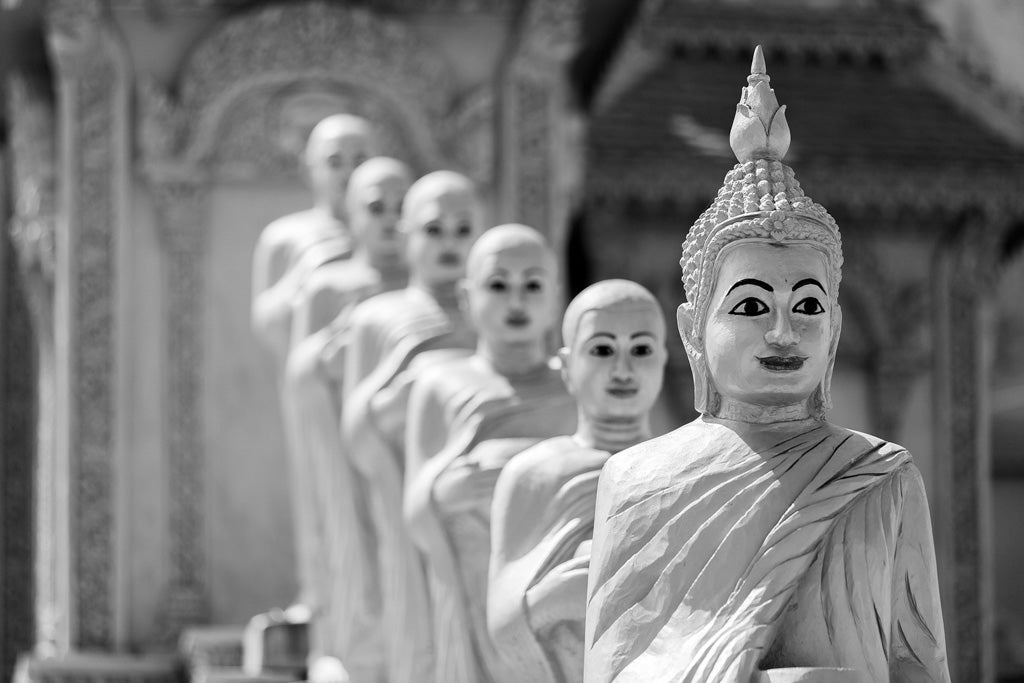 A black and white image titled "Temple Guardians" by FN Prints displays a row of intricately carved stone Buddha statues, depicting Buddhist monks in robes. The front statue features detailed facial features and a small topknot. Ornately decorated Cambodian temple architecture enhances the spiritual imagery in the background.