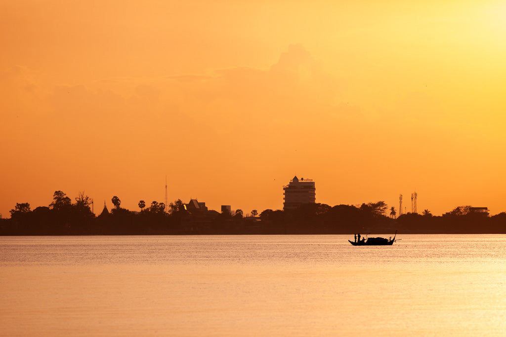 A small boat floats on calm water at sunset, with a vibrant orange sky and a silhouetted cityscape in the background. FN Prints' "Golden Tranquility" photograph beautifully captures this serene scene, showcasing distant buildings and trees along the shoreline reminiscent of Cambodian river cultural landscapes.