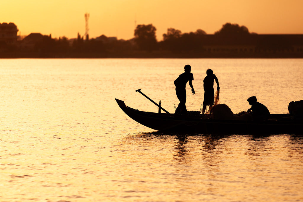 Lit by the Cambodian river's golden sunset, silhouetted figures aboard a fishing boat capture a moment of serene beauty in FN Prints' "Golden Fishermen." One person stands tall while another bends down, both reflected in the calm water. The distant shoreline and cultural richness of the background complete this captivating scene.