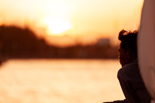 A person is leaning on a railing, gazing at a sunset over a tranquil riverside bathed in warm, golden light, with the water mirroring the vibrant hues of the sky. The background blur highlights the serene and contemplative mood, perfectly capturing the essence of FN Prints' "Moment Of Contemplation.