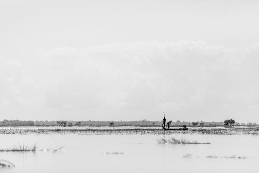 Harvest Waters" by FN Prints is a black and white print depicting two individuals standing in a small boat on a vast, shallow body of water. One person holds a long pole, possibly navigating the boat. The background showcases a distant horizon and scattered vegetation, evoking the serenity of rice field photography.