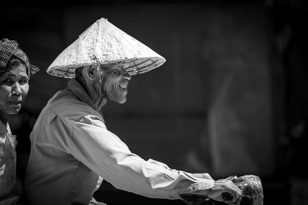 A black and white photo titled "Faces of Joy and Reflection," from FN Prints, captures an elderly person wearing a traditional conical hat while riding a bicycle. Another individual with a similar head covering is partially visible beside them. The blurred background heightens the emotive storytelling, making it perfect for wall art.