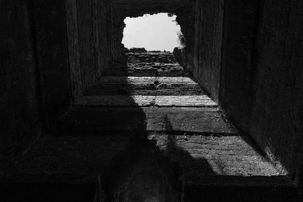 FN Prints' "Ancient Chimney Chute: Angkor Wat" captures a mesmerizing black and white photo from inside an ancient chimney. Sunlight streams through a hole at the top, illuminating the textured, rough walls that cast dramatic shadows and evoke architectural elegance. The stark contrast between the light above and the darkness inside results in a striking visual effect.