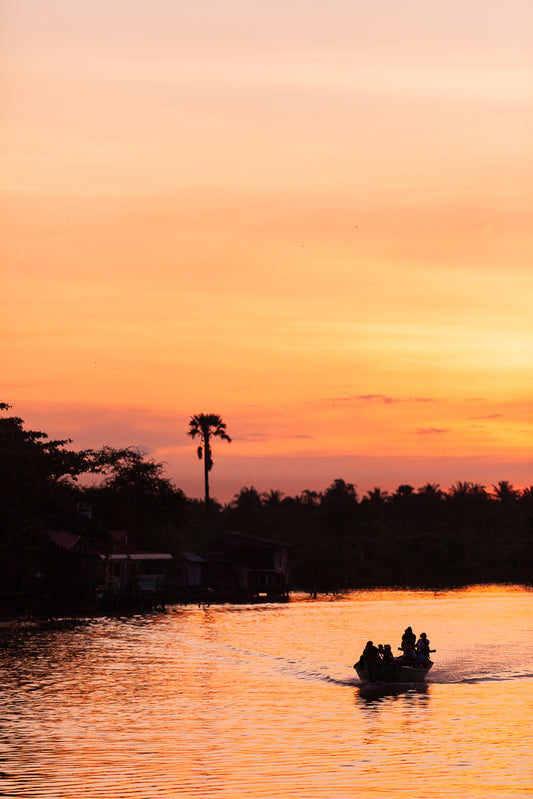 A serene sunset over a calm river with an orange and pink sky. A motorboat with several people aboard sails through the water towards the horizon. Silhouettes of palm trees and houses line the riverbank, creating a scene reminiscent of FN Prints' "Cambodian River at Sunset" wall art.