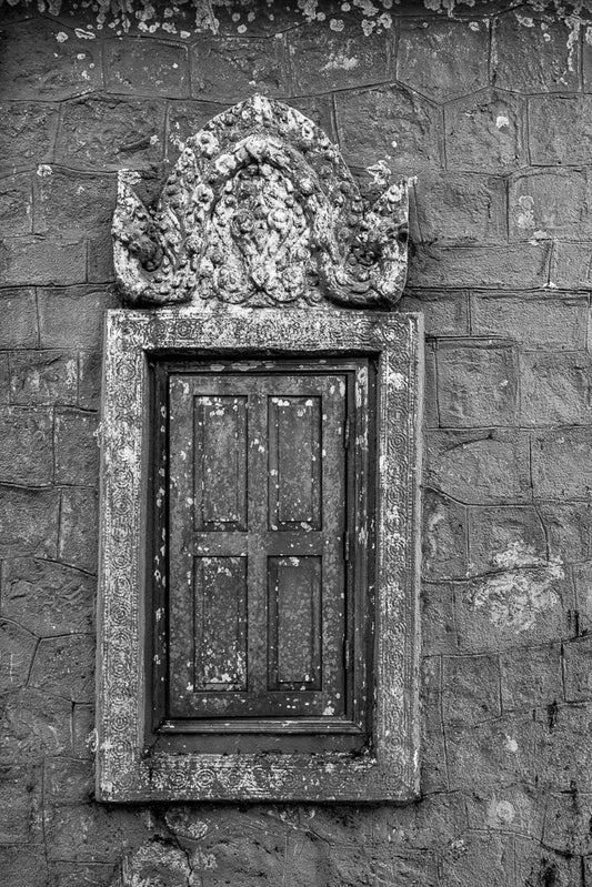 A black and white image from FN Prints' "Window Framed" series captures the historical elegance of an old, closed wooden window on a brick wall. The ornate frame, adorned with weathered stone carvings at the top, features intricate patterns and a central face with a serene expression. The wall shows signs of age and decay.