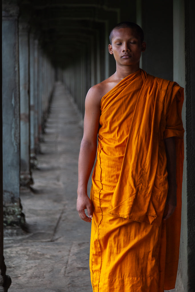 A Cambodian Buddhist monk dressed in an orange robe stands in a serene pose with eyes closed in a narrow, dimly lit corridor with columns lining both sides. The corridor stretches into the distance, creating a sense of infinite depth and spiritual introspection. This captivating scene is beautifully captured in FN Prints' "Eyes Wide Shut.