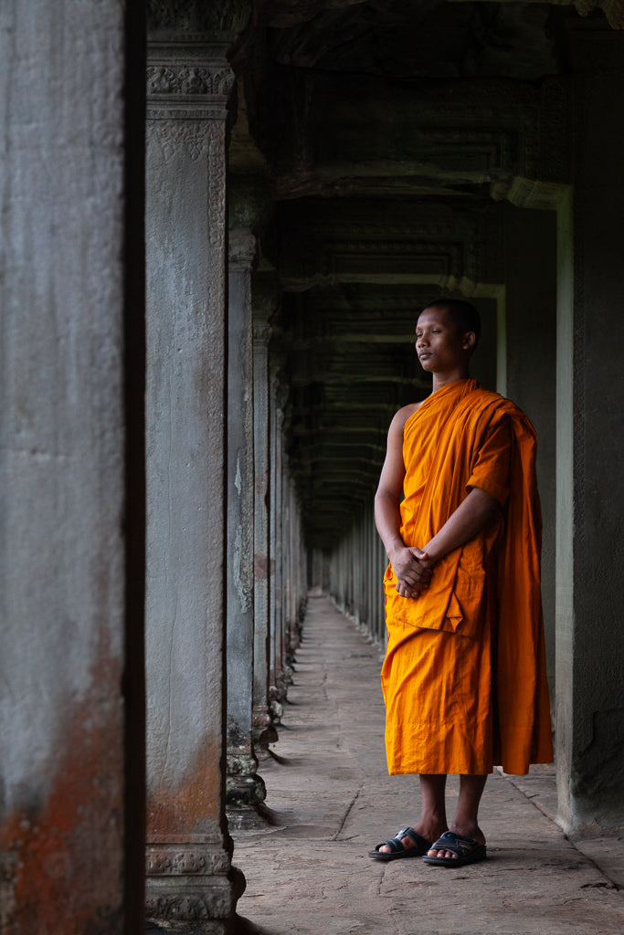 The "Sacred Perspective" wall art piece by FN Prints depicts a person in an orange robe, standing in contemplation within a stone corridor adorned with ornate columns. The individual, evoking the essence of a Buddhist monk photography piece, faces slightly to the left, hands clasped and deep in thought, creating a serene and spiritual ambiance set in an ancient environment.