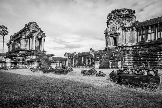 Black and white photography capturing the ancient stone ruins with elaborately carved structures, weathered walls, and remnants of buildings. The scene, reminiscent of FN Prints' "North Thousand God Library: Angkor Wat," shows overgrown grass and scattered stones, suggesting the historical significance and age of the site.