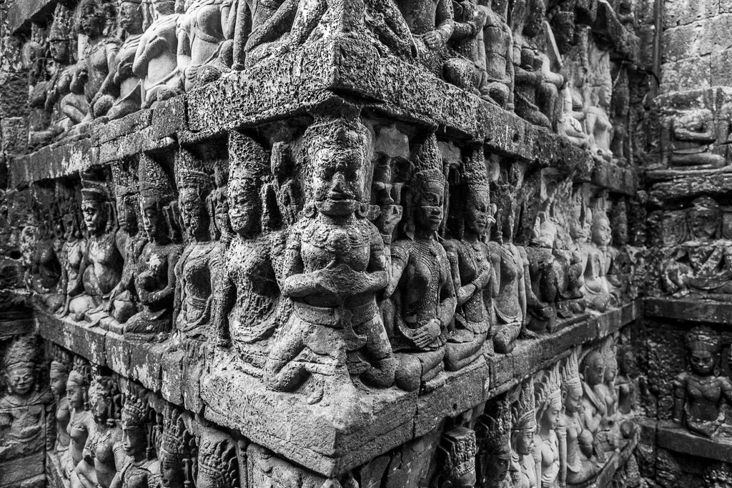 A close-up of elaborately carved stone figures on the walls of an ancient temple at Angkor Thom. The figures, reminiscent of those in "The Terrace of the Leper King: Angkor Thom" by FN Prints, depict various deities and mythical beings in intricate detail. The black and white photography highlights their texture and craftsmanship.