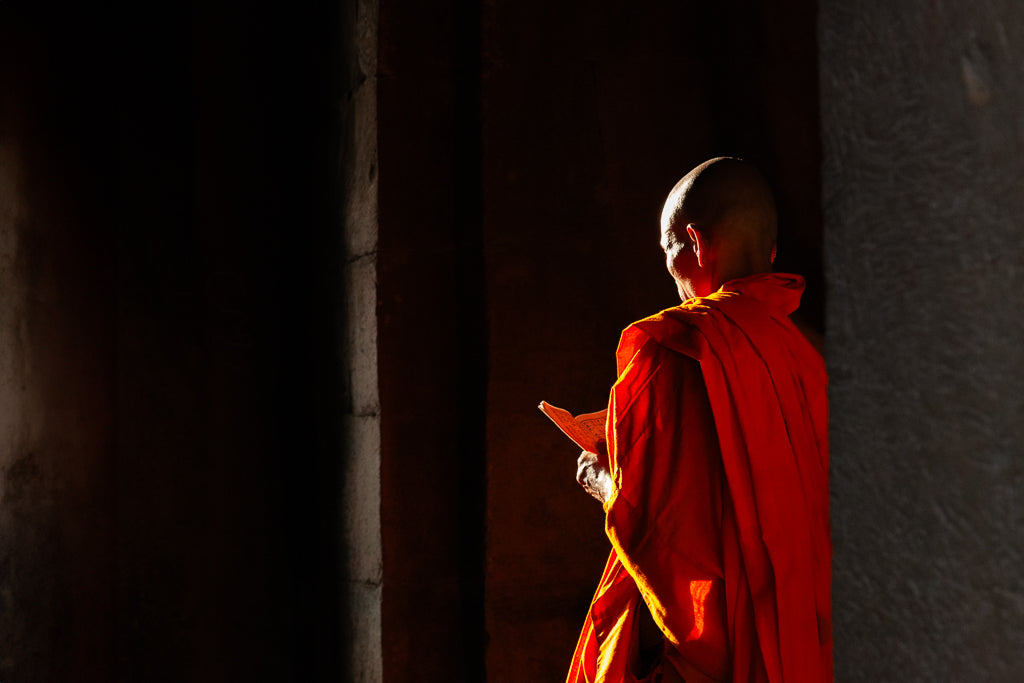 In the serene, dimly lit space, a Cambodian Buddhist monk draped in an orange robe stands with his back to the camera, holding an orange book. Sunlight streams in, illuminating the vibrant color of his robe and evoking a moment of spiritual enlightenment. This tranquil scene is captured exquisitely in "Bathed in Enlightenment" by FN Prints.