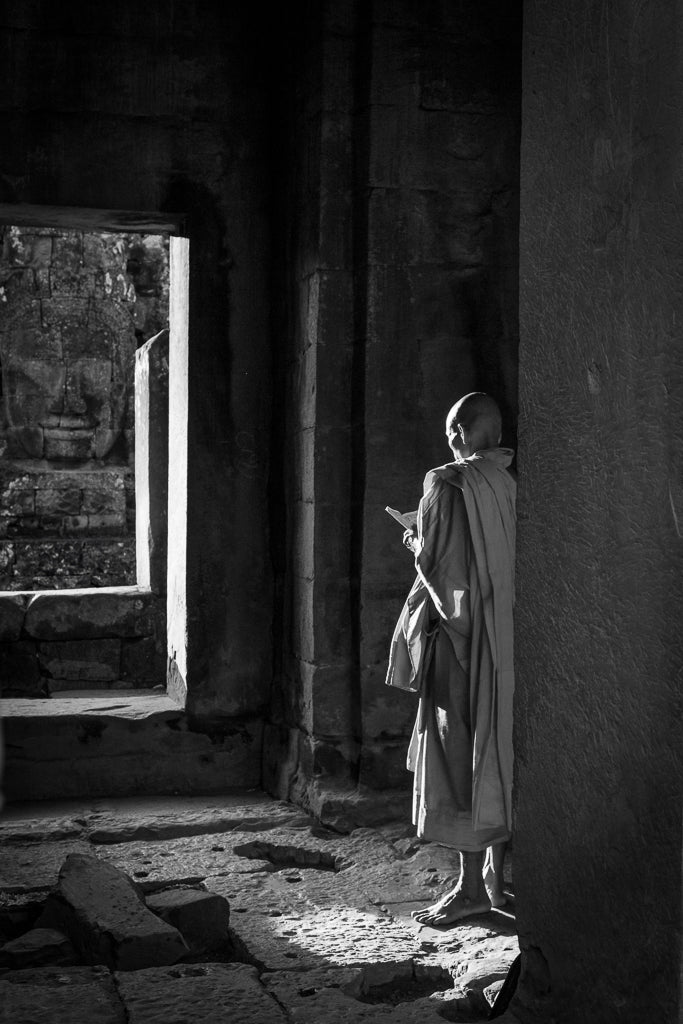 Illuminated Wisdom by FN Prints is a striking piece of black and white photography that captures a Buddhist monk in traditional robes standing beside a large doorway in an ancient temple. Bathed in sunlight, he appears to be meditating or reading. In the background, a large stone face is partially visible through the doorway, creating an impactful piece of spiritual wall art.