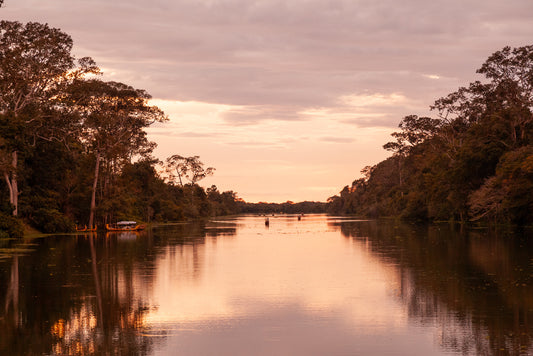 The Sundown River photography print by FN Prints captures a serene Cambodian river scene at sunset, with calm waters reflecting the orange and pink hues of the sky. Flanked by tall trees on both sides, a small boat is moored to the left shore while a lone figure floats in the distance at the center of the river. This idyllic moment is perfectly preserved in this beautiful print.