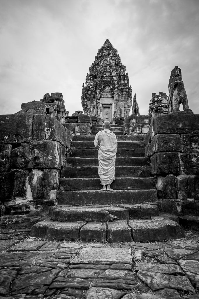 A person dressed in traditional robes ascends the worn stone steps leading up to an ancient temple, reminiscent of Angkor Wat. The intricate details of the architecture are visible, and the overcast sky casts a somber yet serene atmosphere, perfect for spiritual reflection. This scene is beautifully captured in FN Prints' "Journey to the Past.