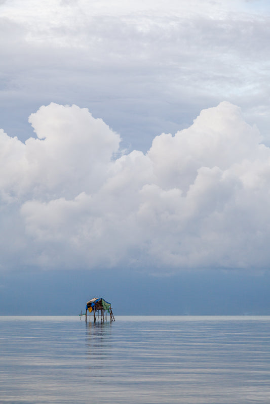 The FN Prints "Vietnamese Stilt Home" captures a solitary stilt house standing serenely in the calm, reflective waters of the ocean. Under a vast sky filled with large, fluffy clouds, this minimalist photography print perfectly showcases the fragile and isolated structure amidst an expansive seascape and dramatic sky, making it an ideal piece of wall art.