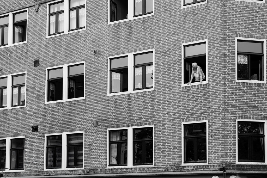 Window Watcher" by FN Prints is a black and white photograph depicting an Amsterdam apartment building with several windows. An elderly person leans out of one window, possibly looking or waving outside. Some windows are open, revealing curtains inside. The scene is calm, capturing a moment of everyday life and architectural symmetry.