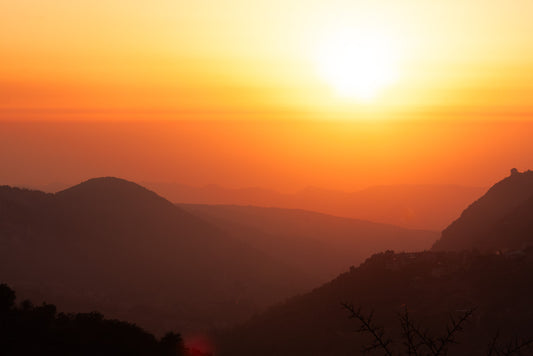 A mesmerizing scene captured by FN Prints' "Mountain Sunset." This artwork showcases a series of mountain ranges bathed in the warm hues of orange and yellow. The sky casts a soft glow over the landscape, with silhouetted hills and serene, fading light creating a peaceful and picturesque mountain vista perfect for any sunset photograph lover.