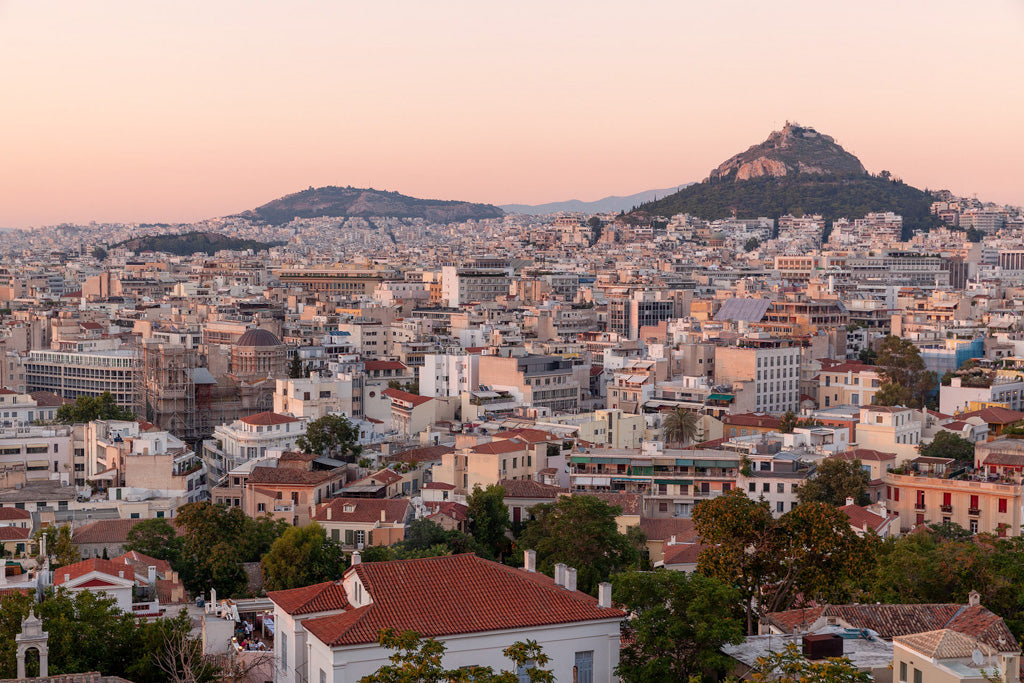 FN Prints' *Athens* captures a breathtaking panoramic view of the Athens cityscape at sunset, showcasing numerous white and beige buildings and rooftops. Two prominent hills rise in the background, one adorned with structures. Mount Lycabettus stands majestically, illuminated by the pinkish hue of golden hour.