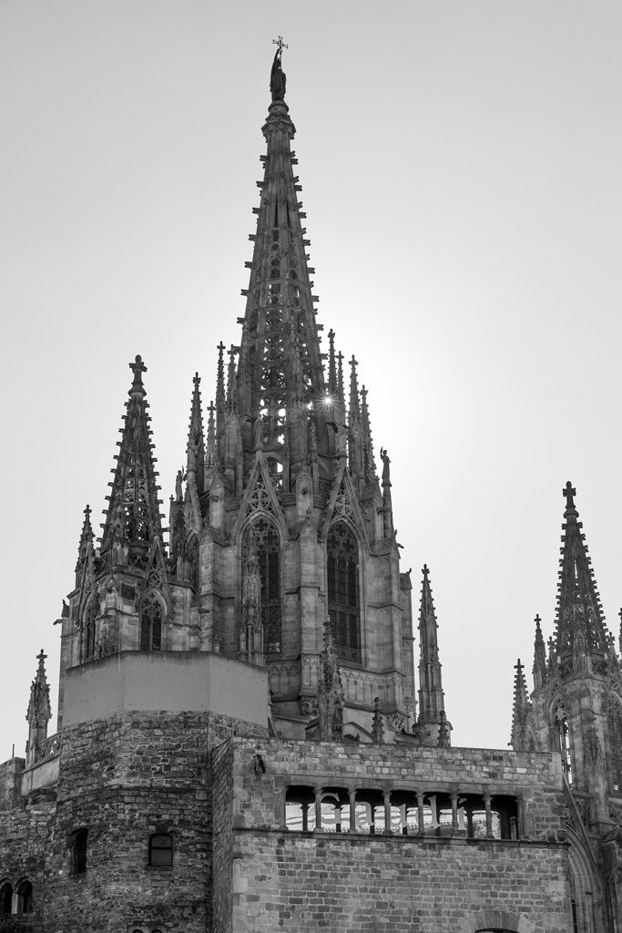 A black and white photograph from FN Prints titled "Barcelona Cathedral Spires" showcases the gothic architecture of the iconic structure, highlighting its intricate spires and detailed stonework. The central spire towers above the building, flanked by two smaller, pointed spires. Arched windows and ornate design elements grace the edifice, with a partially visible wall in the foreground.