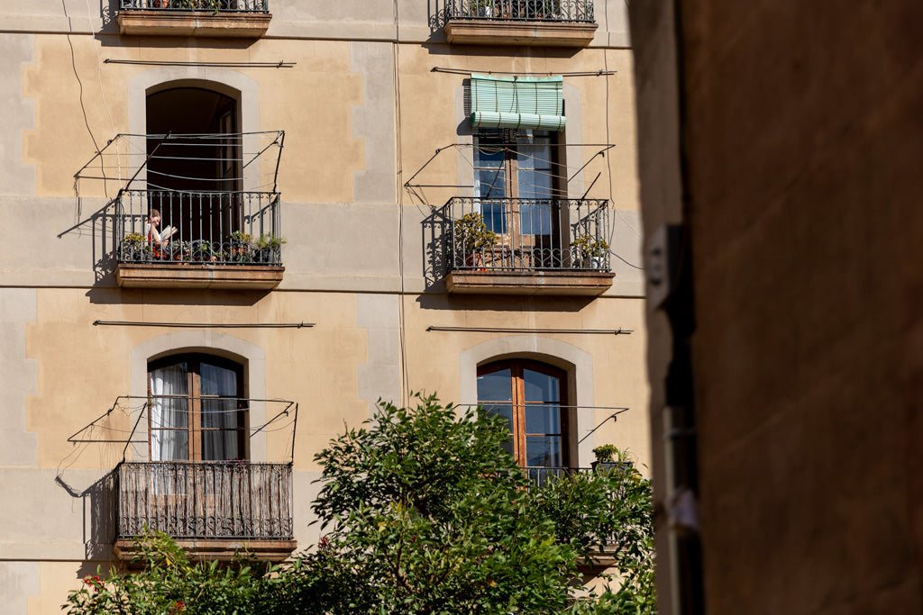 The Reader Colour by FN Prints features a beige apartment building adorned with several small balconies, some embellished with potted plants and green awnings, exuding urban charm. At least one balcony has a person sitting on it. Green trees in the foreground enhance the scene, and a portion of another building is visible on the right side of this vibrant color photograph.