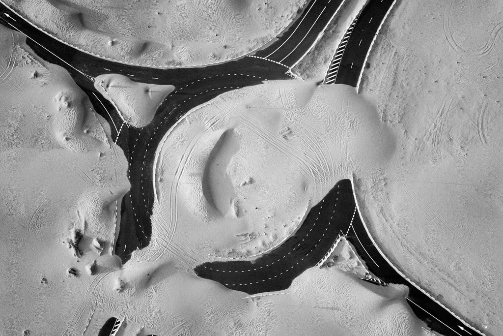 Aerial view of a snow-covered landscape with intersecting roads, forming a complex network of curves and lines against the white snow, much like the FN Prints Desert Roads Roundabout. Tire tracks and subtle shadows add texture to this striking aerial photograph.