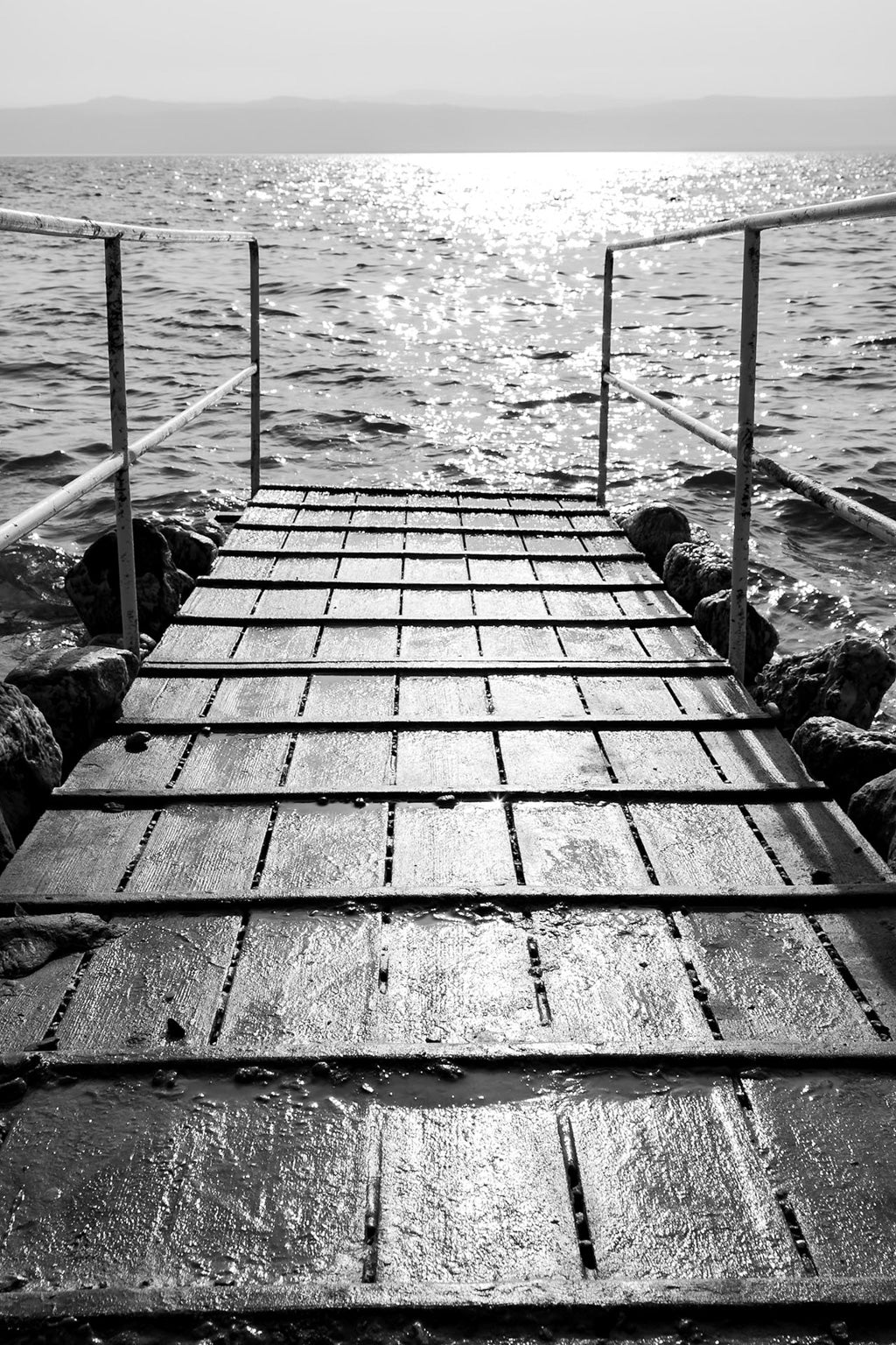 The "Dead Sea Jetty" by FN Prints captures a wooden jetty stretching out into the shimmering sea in a stunning black and white composition. Bathed in bright sunlight, the water sparkles as it flanks the pier, outlined by metal railings and rocky shores. The distant horizon is faintly visible in this serene scene.