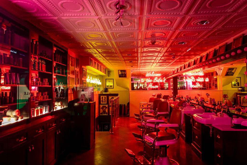 A warmly lit vintage barber shop with red and yellow neon lights casting a colorful glow. The space features retro aesthetics with vintage barber chairs, mirrors, and shelves stocked with grooming products. The ceiling boasts intricate designs, and the neon sign reads "Welcome To The Baba's House," a touch of color from FN Prints.