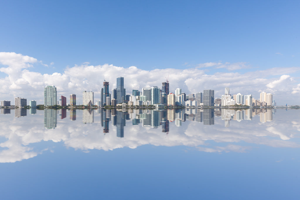 Experience the "Miami Skyline" by FN Prints, featuring a breathtaking panoramic view of Miami's modern skyscrapers under a clear blue sky. The serene, mirror-like water below perfectly reflects the skyline, while fluffy white clouds add texture and enhance the symmetry. Ideal for urban photography enthusiasts or as stunning coastal wall art.

